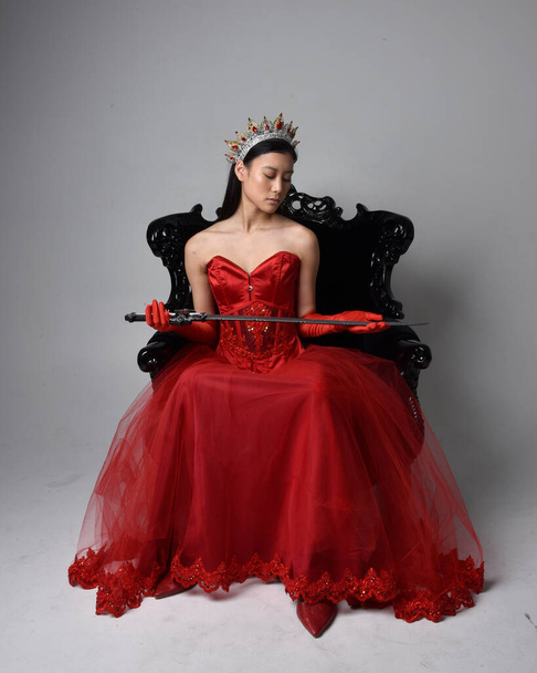 Full length  portrait of beautiful young asian woman wearing red corset, long opera gloves and ornate gothic queen crown. Graceful posing while seated on chair, isolated on studio background. - Foto, Bild