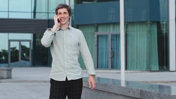 Happy young man walking city street alone holding phone smiling talking with his friend, girlfriend or parents. Millennial guy enjoys free time, quietly spends weekend, relaxes on Friday, makes date - Footage, Video