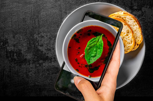 woman hand holding and showing smart phone takes a photo tomato soup with basil in a bowl on Dark grey black slate background. food photos - Foto, Imagem