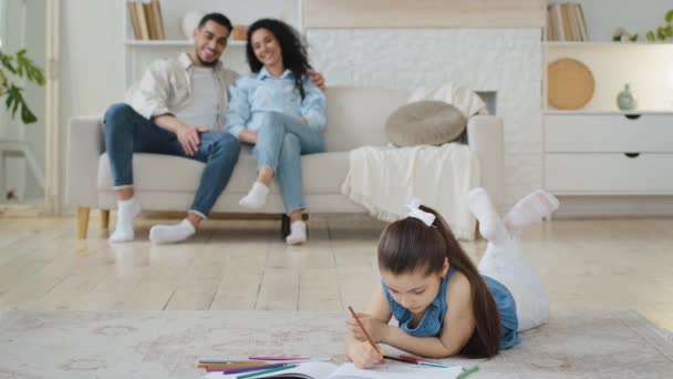 Dcera školačka dítě dívka leží na podlaze kreslí obrázek na papíře s barevnými tužkami, zatímco mladí španělští rodiče sedí na pohovce v obývacím pokoji mluví, rodina tráví čas spolu doma - Záběry, video