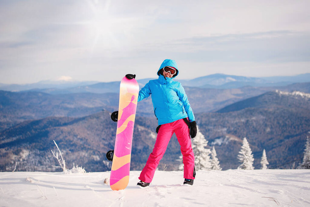Femme snowboarder en vêtements de ski pose avec une planche sur un fond de montagnes bleues. Veste à capuche, pantalon rouge. Mode de vie sain. Concept sportif. Concentration sélective. - Photo, image