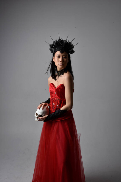 portrét krásné mladé asijské ženy v červeném korzetu a koruně, pózující v gotickém hororovém stylu s kreativními gesty rukou izolovaných proti tmavému černému pozadí studia. - Fotografie, Obrázek