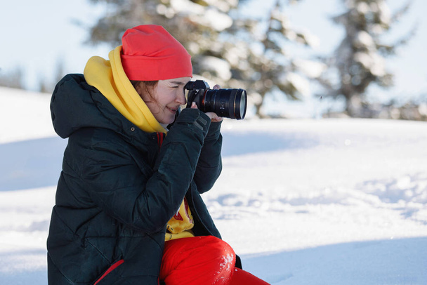 Női fotós a sífelszerelés készít képeket ül a hóban. Egy kamerát tart hatalmas lencsével. Téli ruházat: nadrág és meleg kabát, kötött sapka, síszemüveg. Szelektív fókusz. - Fotó, kép