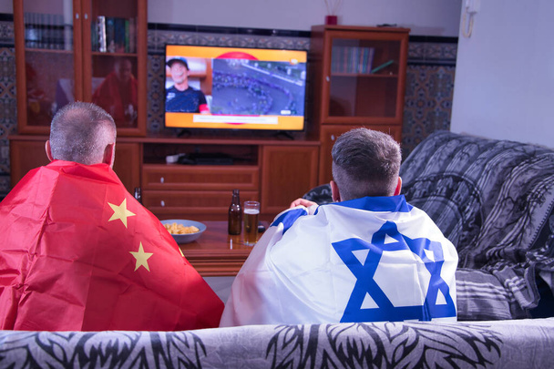 Dwóch mężczyzn, jeden dojrzały i jeden młody oglądający zawody sportowe w telewizji z flagami Chin i Izraela. - Zdjęcie, obraz