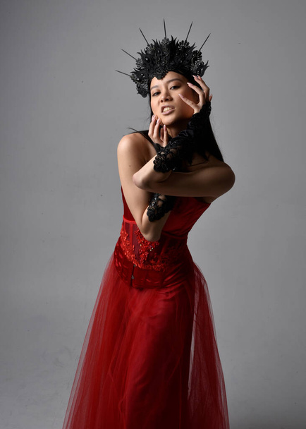 portret pięknej młodej azjatyckiej kobiety ubranej w czerwony gorset i koronę, pozującej w gotyckim stylu horroru z twórczymi gestami dłoni wyizolowanymi na ciemnym czarnym tle studia. - Zdjęcie, obraz