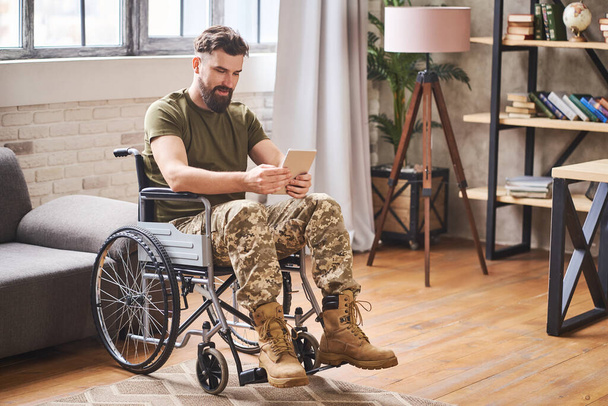 Disabled handsome military man sitting in a wheelchair and using a tablet - Фото, изображение
