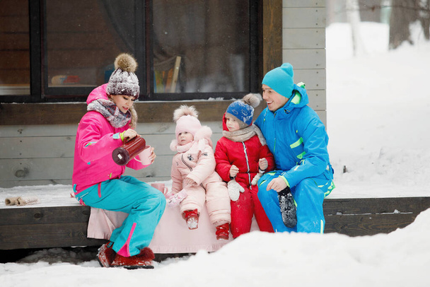 A család téli ruhában ül a verandán és teát vagy kávét iszik. Vidéki ház egy havas tűlevelű erdőben. Meleg zakók, kötött sapkák, poharak. Családi boldogság, értékek és hagyományok. - Fotó, kép