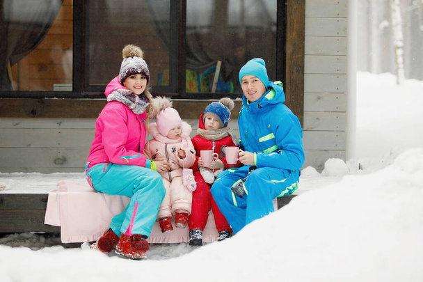 Perhe talvivaatteissa istuu kuistilla ja juo teetä tai kahvia. Maalaistalo lumisessa havumetsässä. Lämpimät takit, neulotut hatut, kupit. Perheen onnellisuus, arvot ja perinteet. - Valokuva, kuva