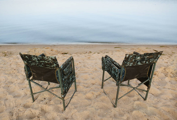 Camouflage fishing chairs on sandy beach near river - Zdjęcie, obraz