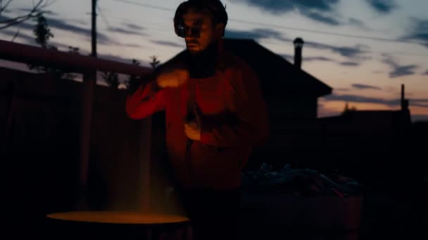 Hombre con gafas y una chaqueta roja tomando el sol cerca de un cubo de basura en llamas en su cara. - Metraje, vídeo