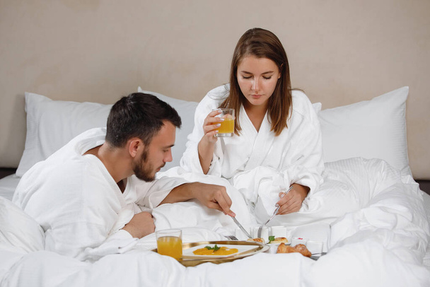 Bearded man and a woman in white coats are having breakfast in bed. Selective focus. - Foto, afbeelding