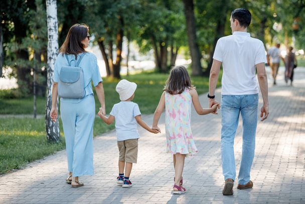 Family couple with daughter and son are walking along the path in the summer park. Happy childhood. Parental care. Back view. Selective focus, blurred background. - Fotografie, Obrázek