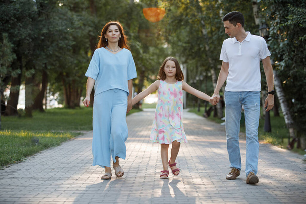 Family couple with daughter are walking along the path in the summer park. Happy childhood. Parental care. Selective focus, blurred background. - Photo, Image