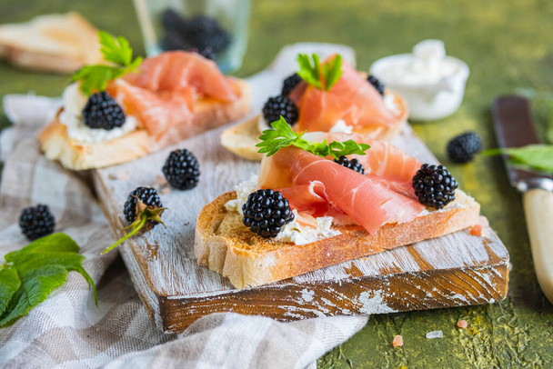 Snack, open sandwich with prosciutto, cream cheese and fresh blackberries on a wooden board on a green concrete background. Sandwich recipes. Italian food - Valokuva, kuva