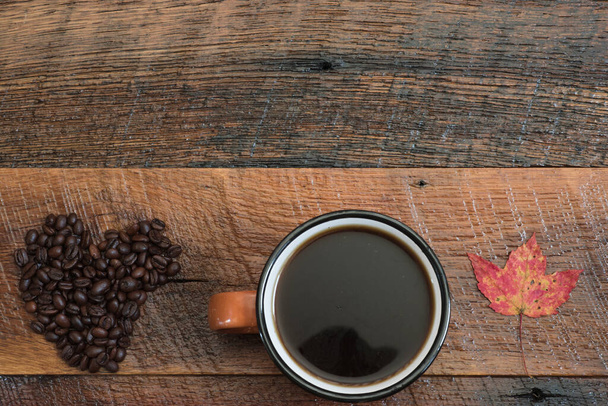 Granos de café en forma de corazón, taza de café caliente y una colorida hoja de otoño en una mesa de madera rústica.  - Foto, Imagen