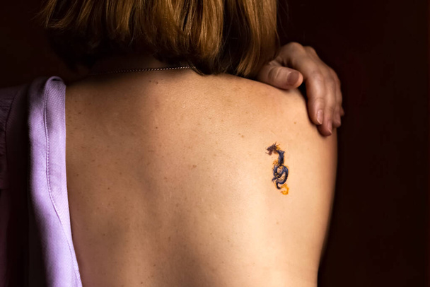La schiena nuda di una giovane donna con i capelli rossi e un tatuaggio sulla spalla. - Foto, immagini