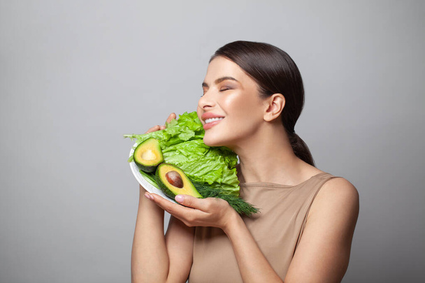 Прекрасная молодая женщина весело улыбается и показывает авокадо и зеленый салат на тарелке. Детоксикация, вегетарианство, здоровое питание и диета - Фото, изображение