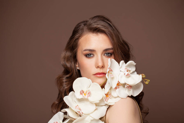 Kıvırcık saçlı, genç ve şık bir kadının güzel moda portresi koyu kahverengi arka planda beyaz çiçekler tutuyor. - Fotoğraf, Görsel