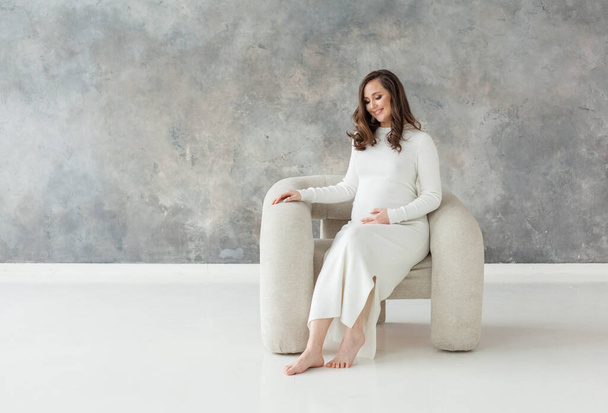 Cute pregnant woman in white dress resting on white armchair on gray banner background - Photo, Image