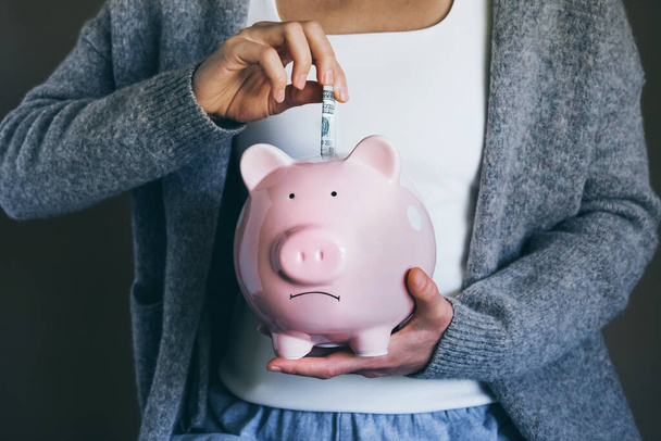 Woman putting bill in sad piggy bank. Female saving money for household payments, bank bills, calculating monthly family budgets, making investments strategy for retirement. Economic crisis concept. - Zdjęcie, obraz