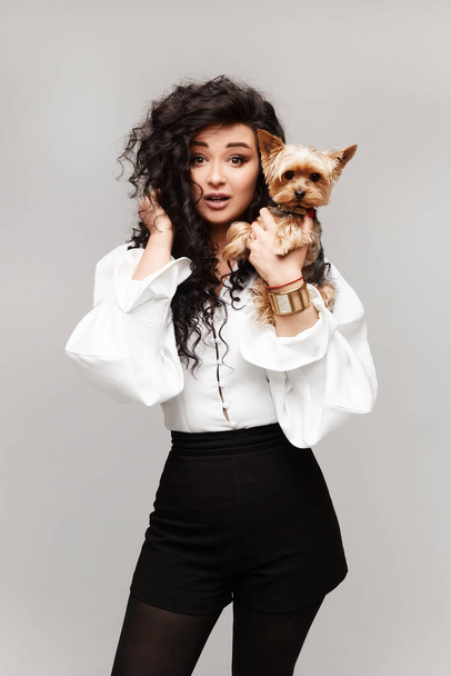 Cropped fashion portrait of a young model woman in a trendy white blouse and black shorts posing with cute Yorkshire terrier over white background, isolated - Photo, image