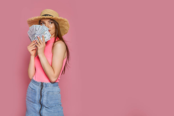 Smiling young tourist girl in straw hat hold fan of cash money in dollar banknotes isolated on pink background. - Фото, зображення