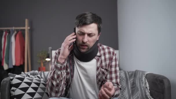 Een man met een schuim halsband die telefoneert, van streek van het nieuws. Man in cervicale brace roept bij de verzekeringsmaatschappij voor medische hulp. Persoon na auto-ongeluk met rugletsel thuis - Video
