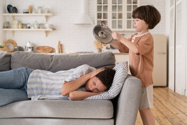 Cansado agotado madre cubre las orejas acostado en el sofá con aburrido hijo ruidoso hacer ruido ruidos utensilios de cocina - Foto, Imagen