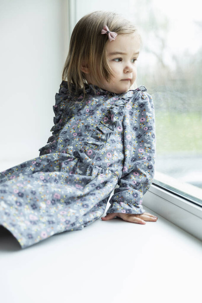 A little girl is sitting by the window. Child in a dress with a floral print. Vertical. - Foto, immagini