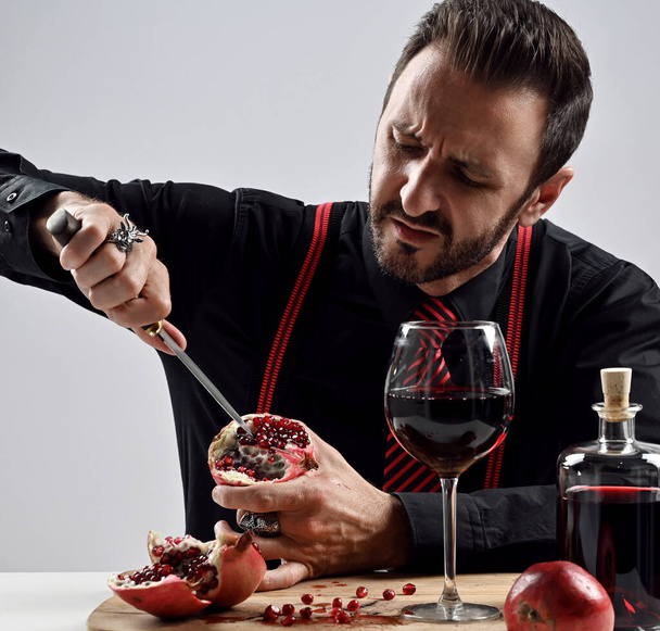 Brutal bearded man in black shirt with striped tie and suspenders at table eats fresh pomegranate sticks knife in it - Фото, зображення