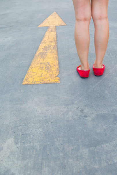 Девушка носить красные ботинки, идущие в направлении с желтой стрелкой движения указатель на асфальтовом фоне дороги - Фото, изображение