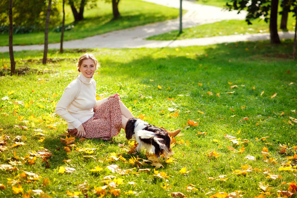 A young blonde woman is sitting on the grass of an autumn sunny park and playing with her Cavalier King Charles Spaniel dog. A woman and her pet in nature. - Photo, Image