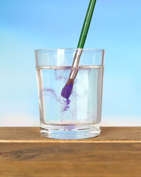 Brush with color paint in glass of water, on wooden table, on blue background - Foto, Imagen