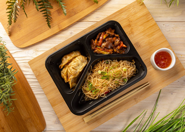 Flat lay of noodles with beef stew and drumplings in take-out box on bamboo chopping board - Zdjęcie, obraz