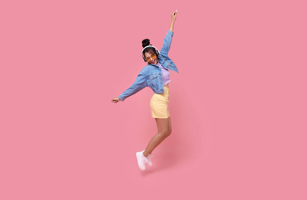 Happy beautiful Asian teen woman in yellow glasses listening music in headphones and dance on pink background.	 - Foto, afbeelding