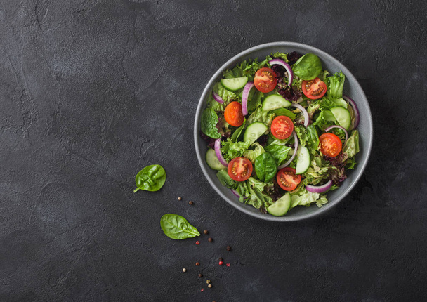 Fresh healthy vegetarian vegetables salad with tomatoes and cucumber, red onion and spinach in grey bowl plate on dark background. Top view. - Photo, Image