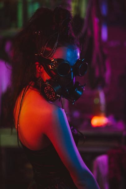 Soldatin im Cyberpunk-Stil mit Gasmaske und Brille mit Neonbeleuchtung in der Garage. Steampunk-Stil - Foto, Bild
