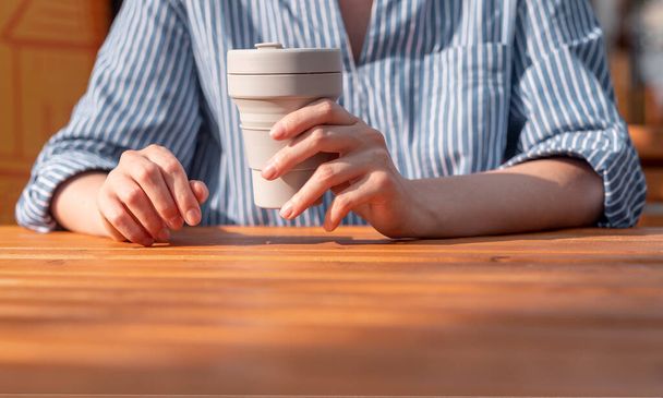 Frau reicht Nahaufnahme mit wiederverwendbarem Silikon zum Mitnehmen Öko-Kaffeetasse in den Händen über Holztisch - Foto, Bild
