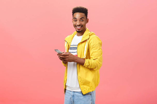 Κομψό ενθουσιασμένοι αφροαμερικανός άνδρας με afro σγουρά κούρεμα στέκεται μισογύρισε πάνω από ροζ τοίχο κρατώντας smartphone φορώντας κίτρινο μοντέρνο σακάκι χαμογελώντας χαρούμενα δείχνει φίλο χαρακτηριστικά της συσκευής - Φωτογραφία, εικόνα