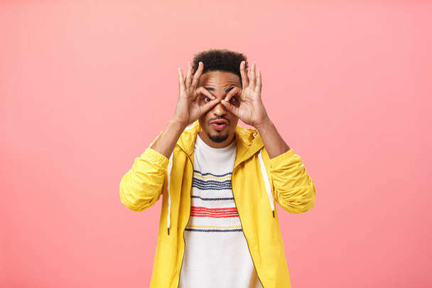 Guy being impressed cannot believe own eyes gasing throught circles made from fingers at camera excited and amazed standing over pink background in trendy yellow jacket intrigued and stunned - Foto, Imagen