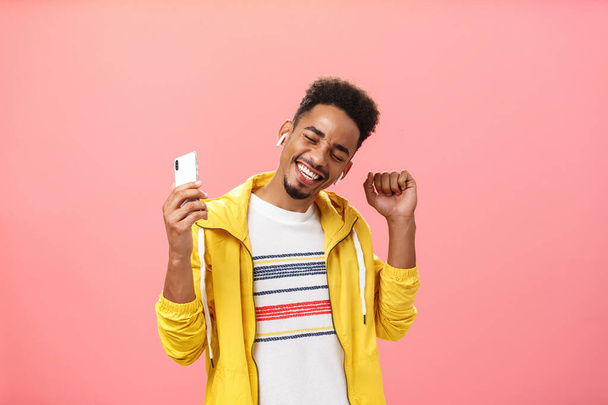 Waist-up shot of stylish carefree optimistic african american male in wireless earphones holding smartphone dancing joyfully with raised hands and broad smile enjoying great music with new headphones - Фото, изображение