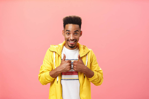 Energético satisfeito e encantado afro-americano macho com barba e penteado afro na elegante jaqueta amarela levantando os polegares em como gesto e sorrindo favorável aprovar ideia legal sobre a parede rosa - Foto, Imagem