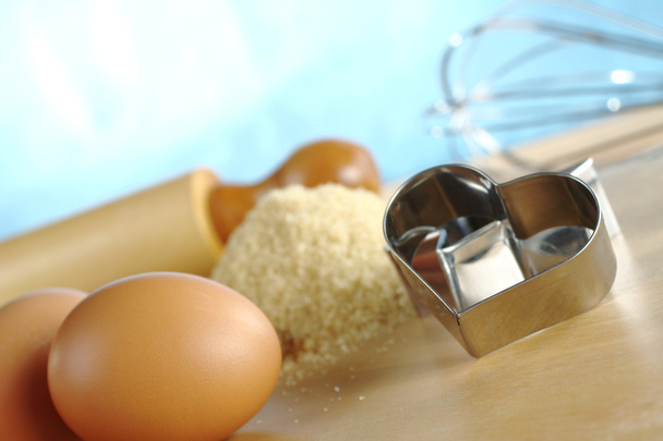 Sydämen muotoinen evästeleikkuri, jossa on leivontaainesosia ja välineitä, kuten munia, ruskeaa sokeria, valssaustappia ja vatkainta (Selektiivinen Focus, Keskity etuosaan
 - Valokuva, kuva