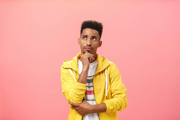Studio shot of concerned thoughtful troubled young dark-skinned guy with beard and afro hairstyle frowning touching chin looking focused at upper left corner making decision over pink background - Photo, Image