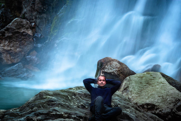 hombre sentado en la roca frente a la cascada corriente de agua blanca que cae de las montañas imagen de larga exposición se toma en la cascada de jang tawang arunachal pradesh. - Foto, Imagen