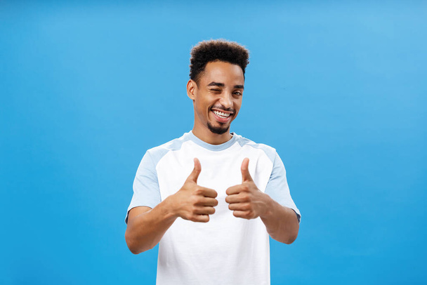 You doing great, support you. Satisfied creative and stylish young african american man with beard winking joyfully smiling broadly showing thumbs up in approval or like gesture over blue wall - Foto, afbeelding