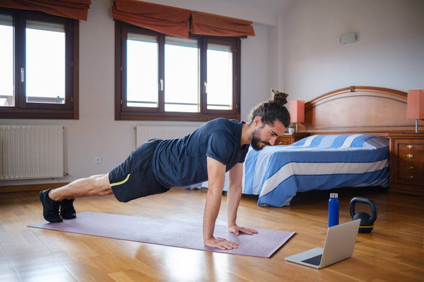 Bärtiger Mann beim Planken, während er zu Hause mit einem Online-Kurs am Laptop trainiert. - Foto, Bild