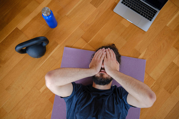 Vyčerpaný muž na podlaze si zakrývá obličej rukama po cvičení doma. - Fotografie, Obrázek