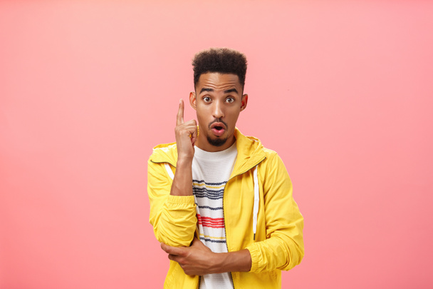 Got it finally understood. Portrait of excited smart dark-skinned creative male with beard in trendy yellow jacket raising index finger in eureka gesture folding lips adding suggestion over pink wall - Valokuva, kuva