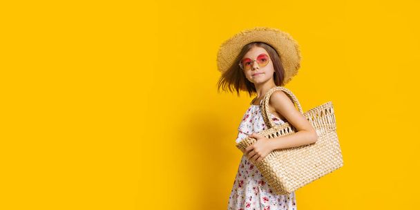 Porträt eines fröhlichen Mädchens mit Sommermütze, Sonnenbrille, Strohsack vor gelbem Hintergrund.  - Foto, Bild